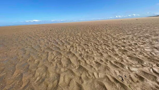 Een breed zandstrand met duidelijke voetsporen en een helderblauwe lucht.