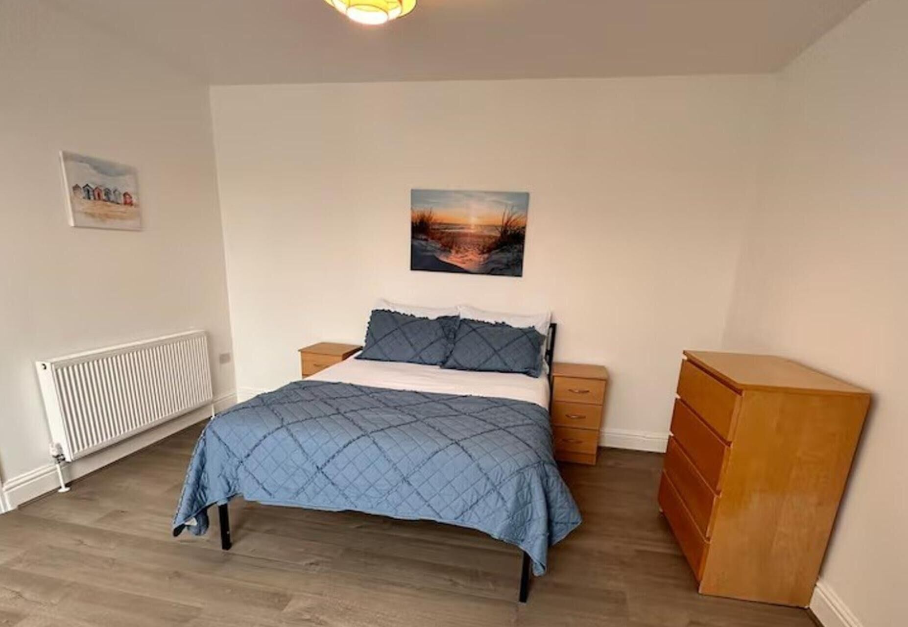 A bedroom with a bed, a nightstand, a dresser, a radiator, and a painting on the wall.