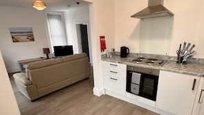 Een kleine keuken met een fornuis, oven en magnetron. In de aangrenzende woonkamer staat een bank en een televisie.