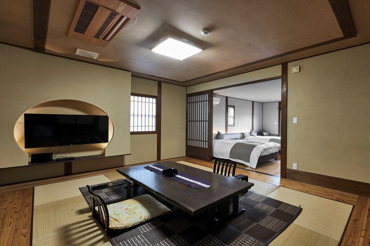 A traditional Japanese-style room with tatami flooring, a low table with cushions, a television, and a sliding door leading to a bedroom.