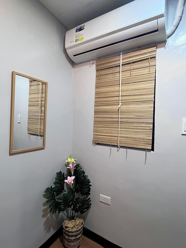 A room with a wall-mounted air conditioner, a mirror, a window with bamboo blinds, and a potted plant.