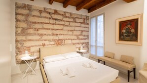 Une chambre avec un mur en briques, un lit recouvert de draps blancs, un banc et un tableau.