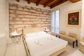 A bedroom with a brick wall, a bed with white linens, a bench, and a painting. - New Bridge House - Domus Verona (Verona)