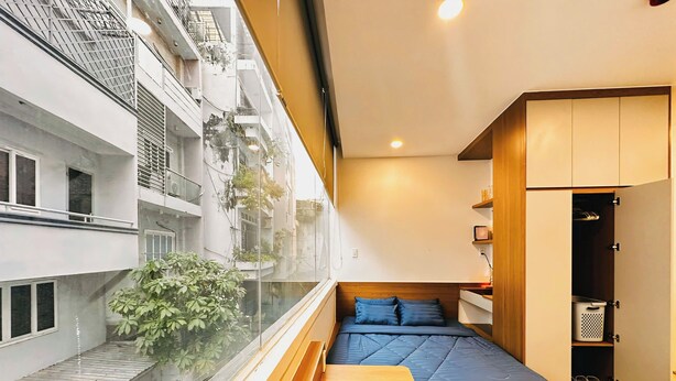 A compact bedroom with a bed, wooden bench, wardrobe, and a view of a neighboring building. - Loftic Apartments - Pink Church (Ho Chi Minh City)