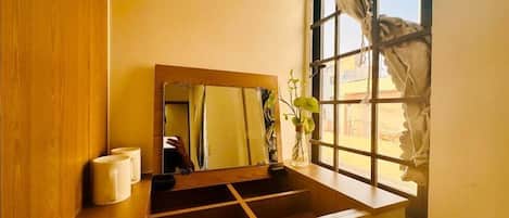 Un baño con tocador de madera, un espejo, un jarrón con flores y una ventana con cortinas.
