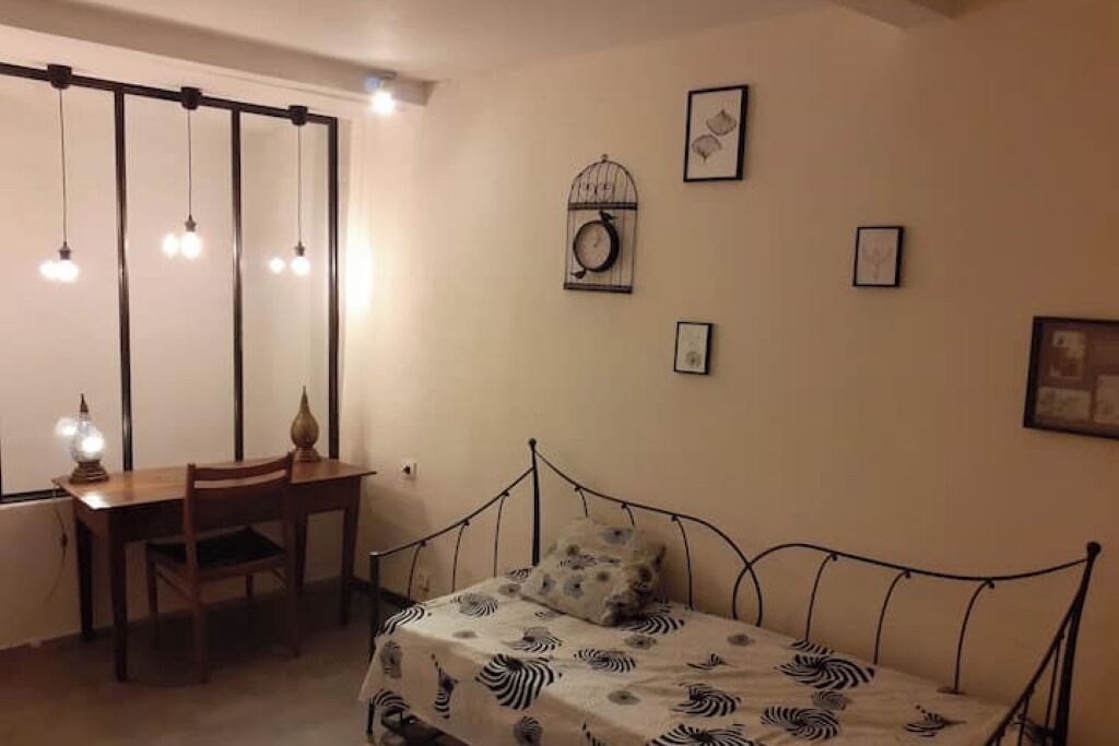 A bedroom with a bed, a desk, and a chair. There are wall-mounted lights and framed pictures.
