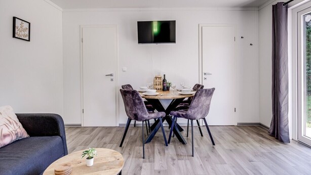 Un moderno salón con zona de comedor, un televisor de pantalla plana en la pared y un amplio ventanal con cortinas.
