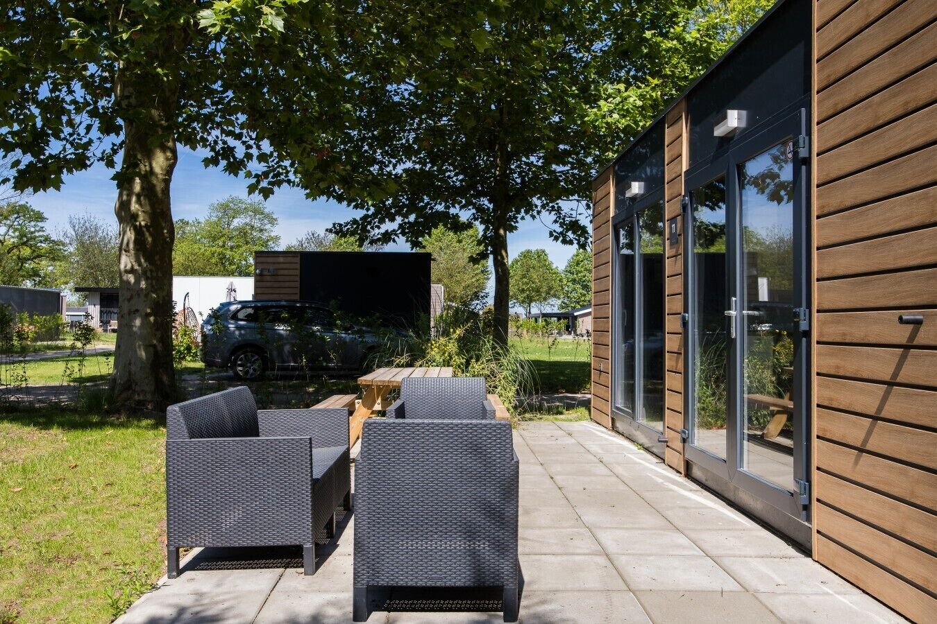 A modern outdoor patio with wicker furniture, a car in the background, and a building with large windows and wooden siding.
