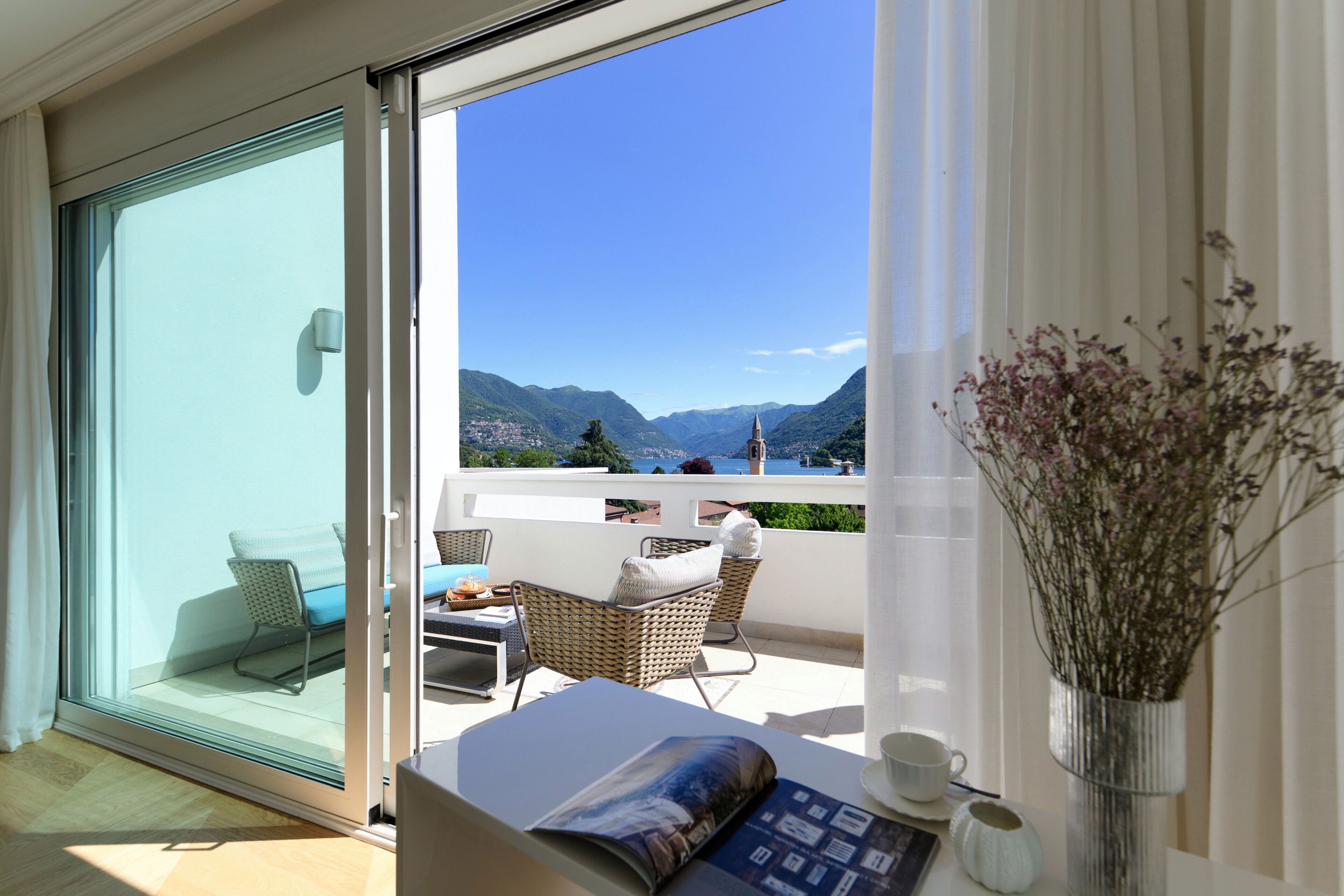 Ein Balkon mit Blick auf Berge, ein Korbstuhl, ein Tisch mit Kissen und eine Vase mit Blumen.