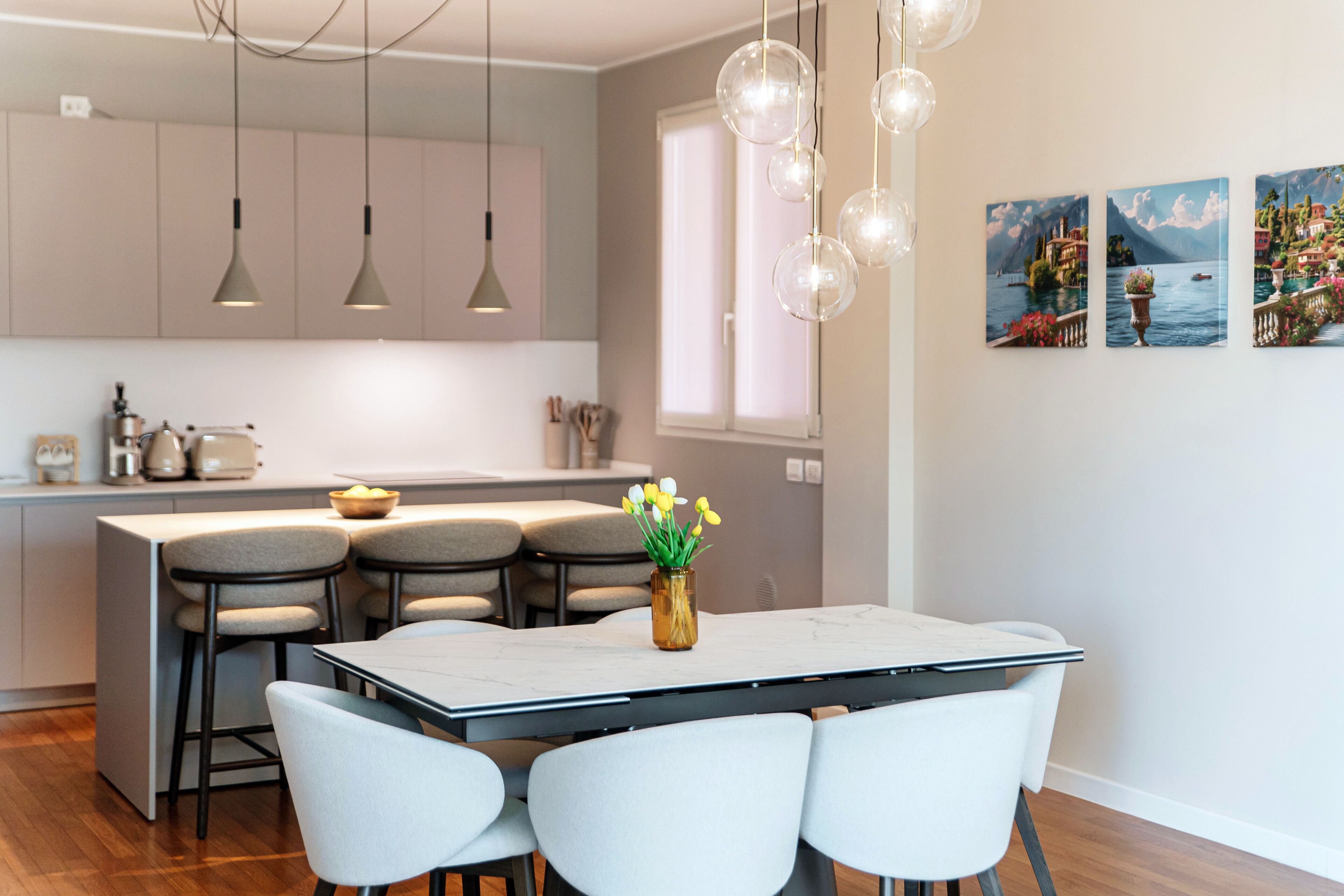 Una cocina moderna con lámparas colgantes, un comedor con mesa y sillas, y un ventanal con vistas a un lago de montaña.