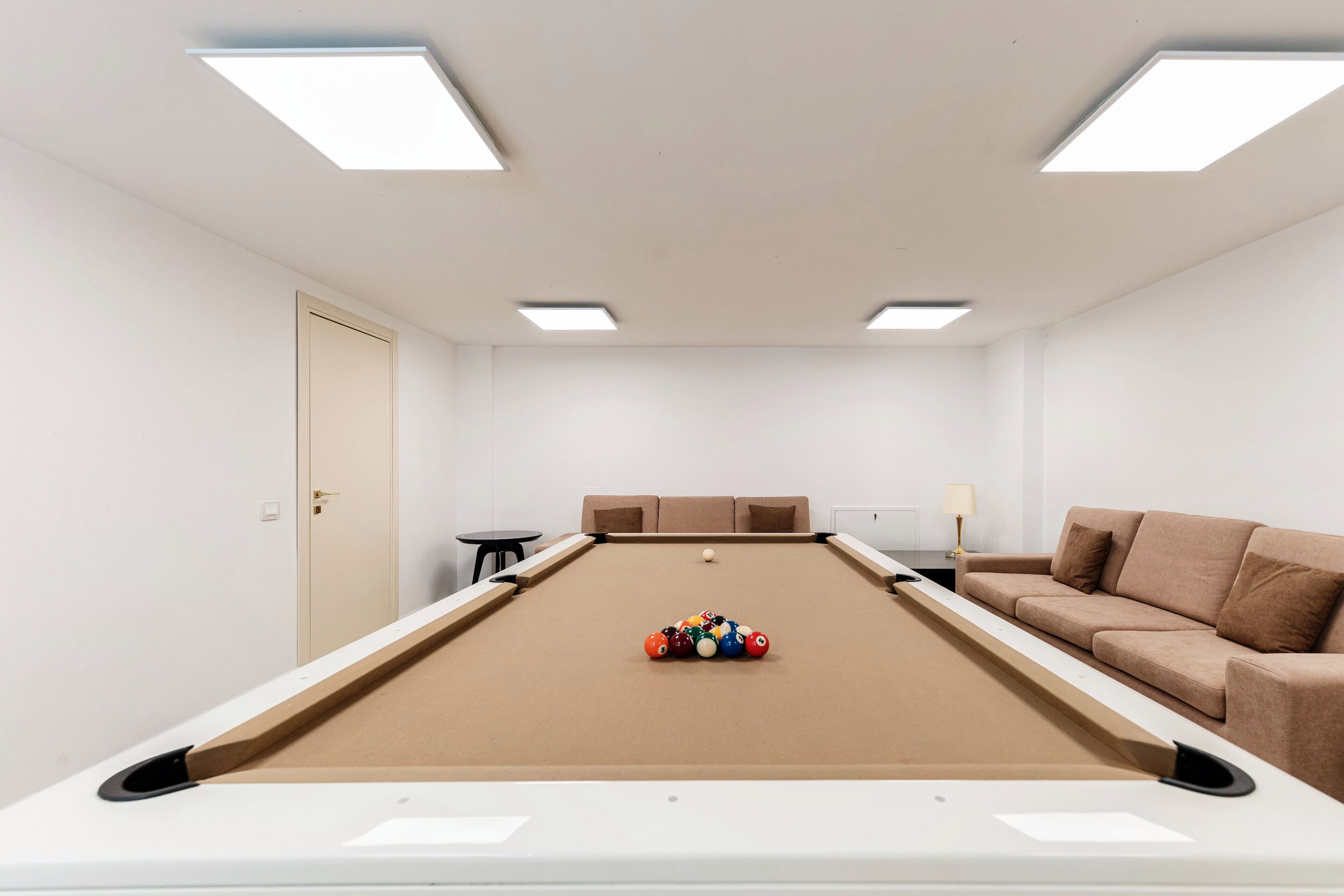 A pool table with balls, a sofa, a lamp, and a door in a room with white walls and ceiling lights.