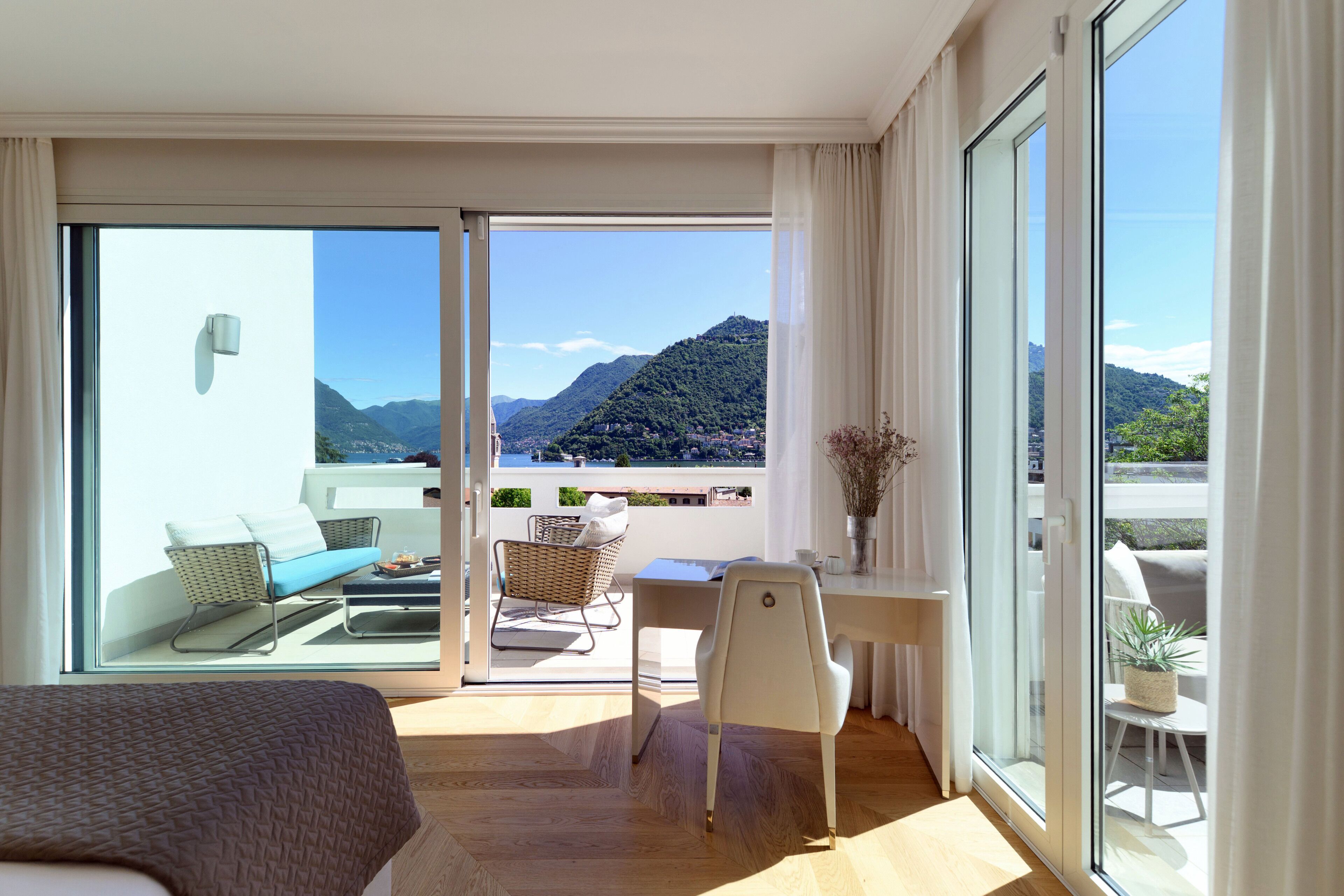 A modern room with a bed, a desk, a chair, and a balcony with a view of mountains.