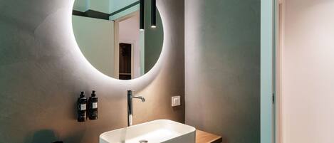 Een moderne badkamer met een ronde spiegel, een wit wastafelmeubel en twee zwarte flessen aan de muur.