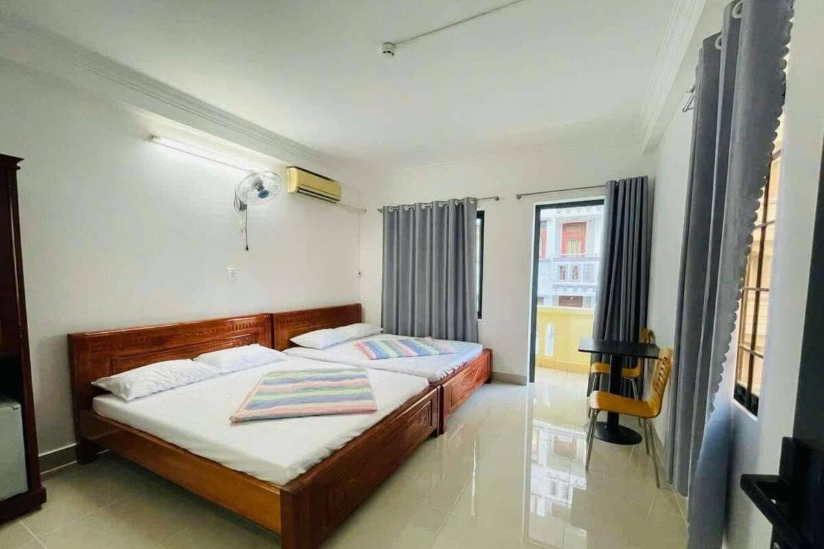 Habitación de hotel con cama de madera, ventilador de techo, unidad de aire acondicionado, puerta corredera al balcón y una pequeña mesa con una silla.