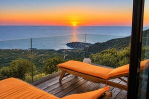 A balcony with a wooden floor and orange cushions, overlooking a coastal landscape with a sunset view. - Harmony House- Sea View Cretan Escape with Jacuzzi (Kissamos)