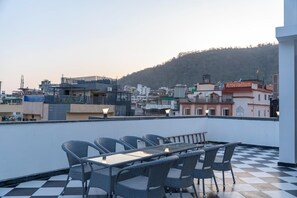 Terrassa al terrat amb sòl a escacs, moble de vimetes i vista a una ciutat amb edificis i un turó.