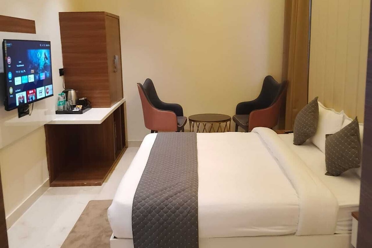 Habitación de hotel con cama, escritorio con computadora, dos sillas, una mesita y televisor en la pared.