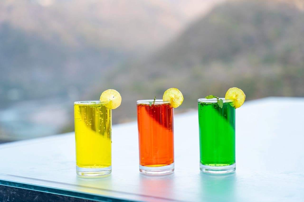 Three glasses of colored liquid on a ledge with a blurred natural background.