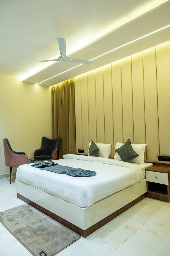 A modern hotel room with a large bed, bedside tables, a ceiling fan, and a seating area with chairs. - Hotel Hrishikesh High Tapovan 2.0 (Rishikesh)
