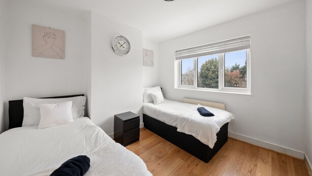 A bedroom with two beds, a nightstand, a wall clock, and a window with a view of trees.