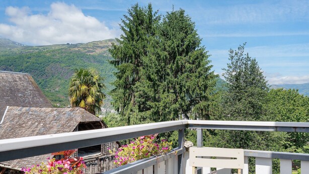En balkon med et bord og stole, der udover et bjergrigt landskab med træer og en bygning.