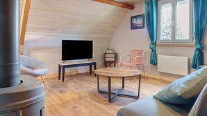 Et hyggeligt værelse med TV, et rundt bord, en sofa, en lænestol, en varmeovn og et vindue med gardiner.