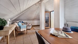 A modern loft bedroom with a sloped ceiling, a wooden dining table, a sofa, and a view through a glass door.