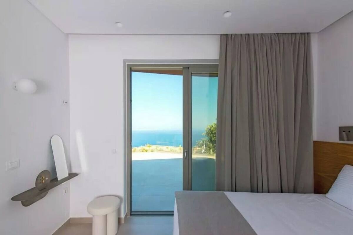 Une chambre d’hôtel moderne équipée d’un lit, d’une petite table de chevet, d’un miroir, d’une fenêtre donnant sur la mer et d’une porte coulissante menant au balcon.