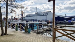 A wooden dock with a large building in the background, a marina with boats, and a clear sky.