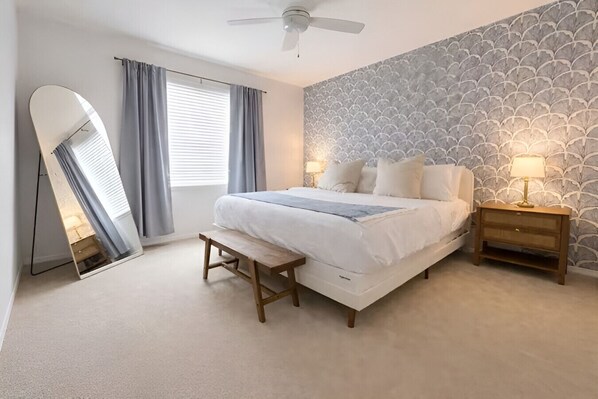 Un dormitorio con cama, mesitas de noche, ventilador de techo, ventana con cortinas y pared con estampado.