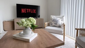 Un soggiorno con una TV a schermo piatto che mostra 'NETFLIX', un vaso bianco con fiori su un tavolino in legno, un divano con cuscini floreali e un mobiletto in legno.