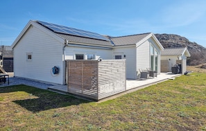 Uma casa de dois andares com telhado solar, uma pequena área de estar ao ar livre e cerca de madeira.