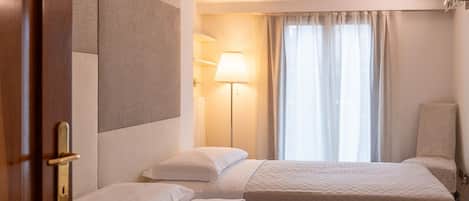 A hotel room with two beds, a lamp, a chair, and a window with curtains.