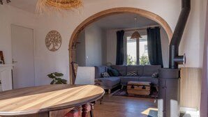 Ein Wohnzimmer mit Esstisch aus Holz, einer Couch, einem Kamin und einem großen Fenster mit Vorhängen.