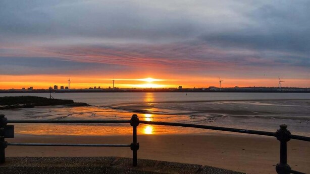 Un atardecer sobre un cuerpo de agua con el perfil de una ciudad a lo lejos y aerogeneradores.
