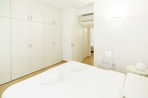 Une chambre d’hôtel minimaliste comprenant un grand placard, un lit avec du linge de maison blanc, une petite table avec une chaise et un appareil de climatisation.