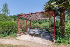Un espace extérieur aménagé avec des sièges, abrité et doté de chaises en métal et d’une table, entouré de verdure et d’un palmier.