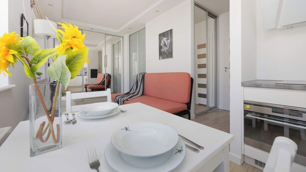 Ein moderner Essbereich mit einem weißen Tisch, gedeckt für zwei Personen, einer Vase mit gelben Blumen und einer Küchenzeile mit eingebautem Backofen.
