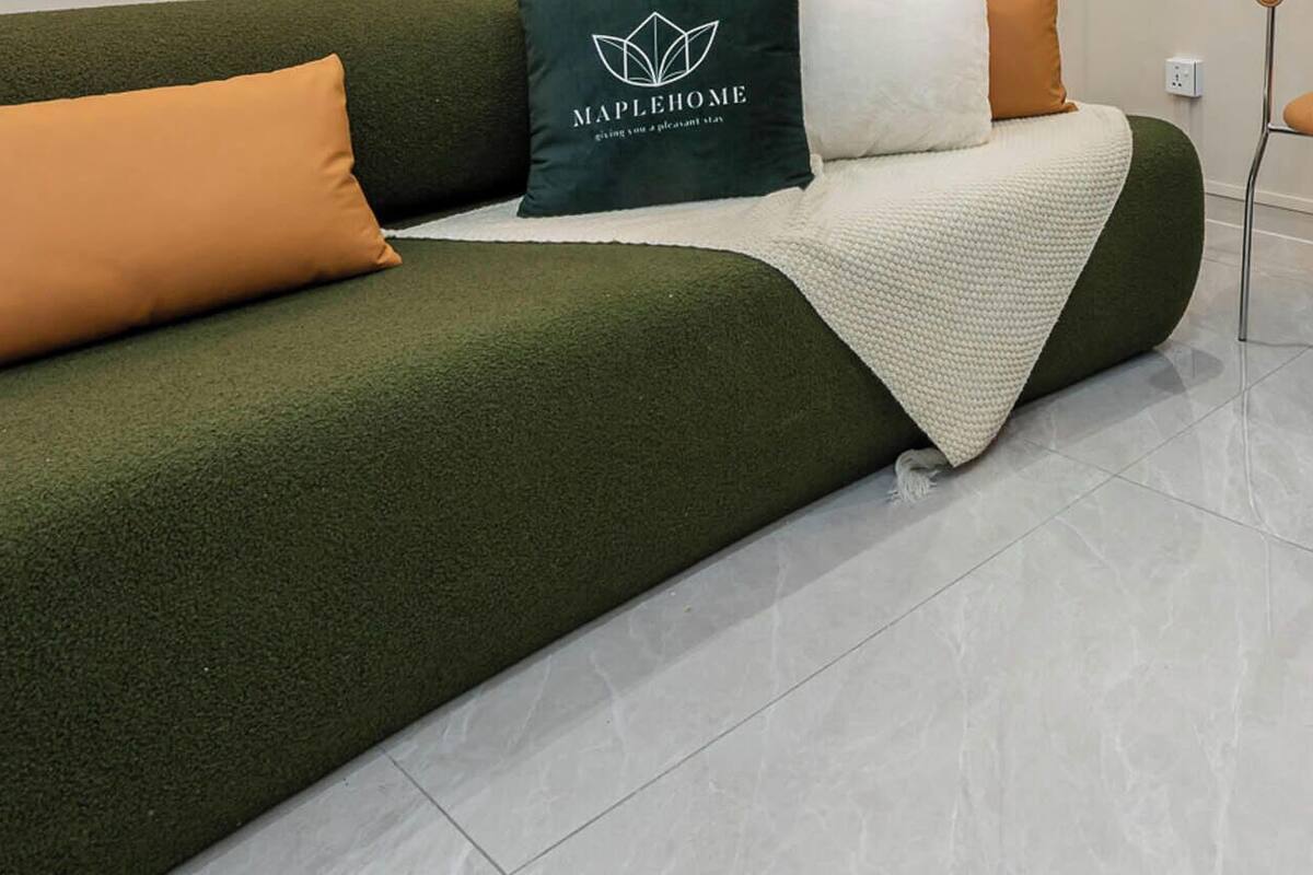 Un sofá verde con una manta blanca y un cojín marrón, una mesita con dos taburetes y un armario de madera oscura.