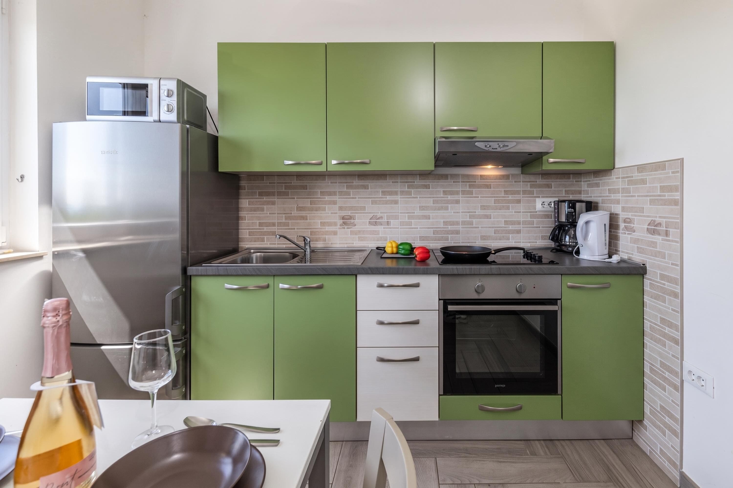 A compact kitchen with green cabinets, a stainless steel refrigerator, a microwave, a stove, and a coffee maker.