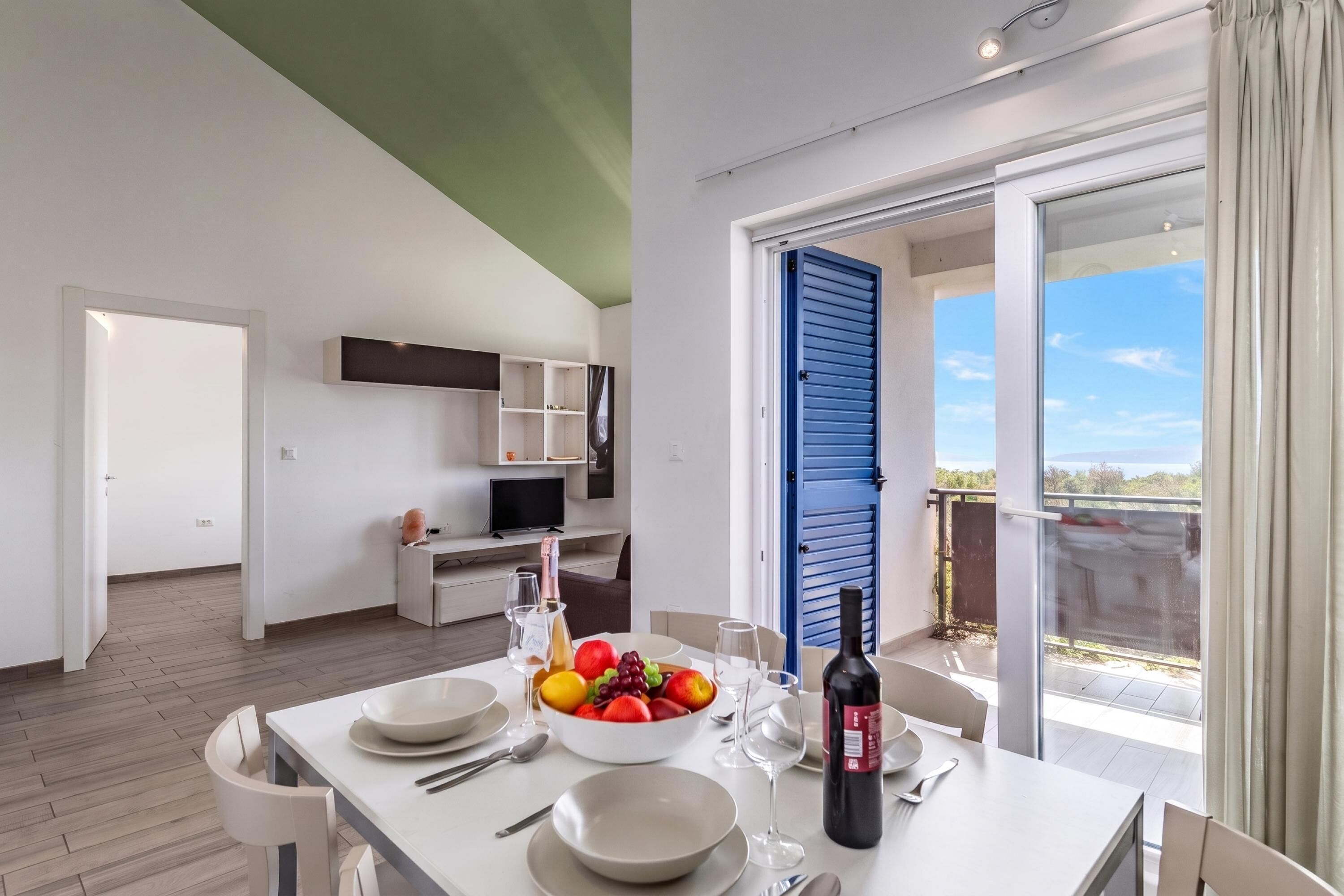 A modern dining area with a table set for two, a television, and a balcony with blue shutters.