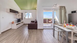 Un espacio habitable moderno con un televisor de pantalla plana, un sofá, una mesa de comedor para dos personas y un ventanal corredero que da a un área exterior.