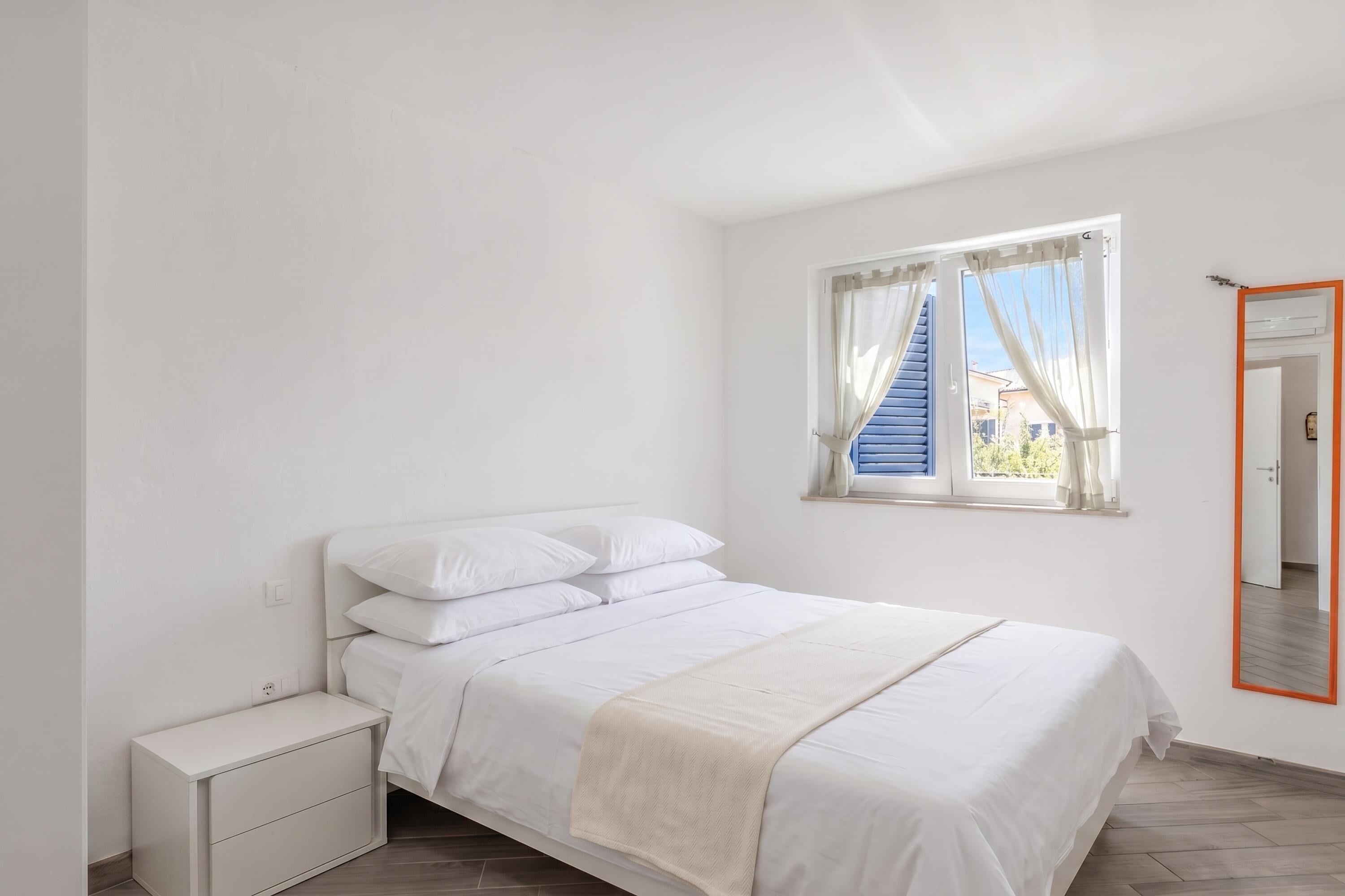 A bedroom with a bed, a bedside table, a window with curtains, and a mirror with an orange frame.