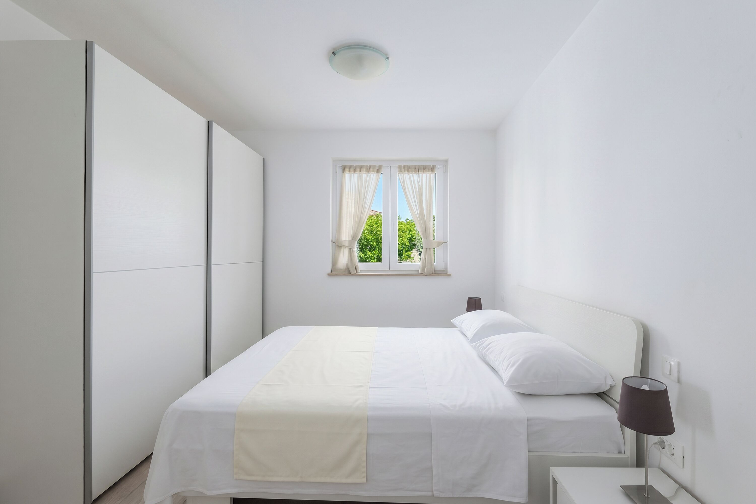 Une chambre à coucher comprenant un lit, une table de chevet, une lampe, une fenêtre avec des rideaux et une armoire.