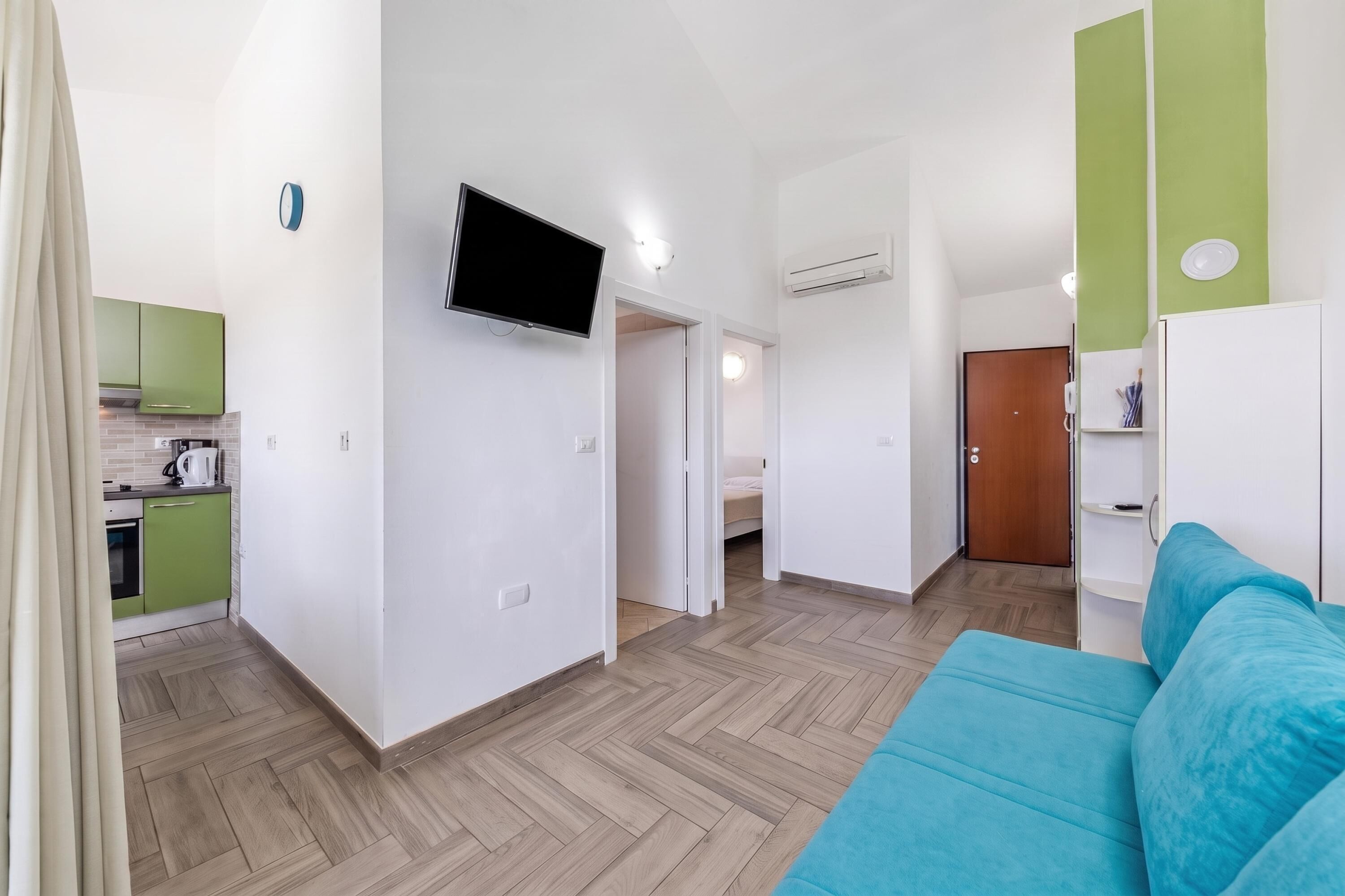 A modern apartment with a kitchenette, a flat-screen TV, a bed, and a blue sofa.