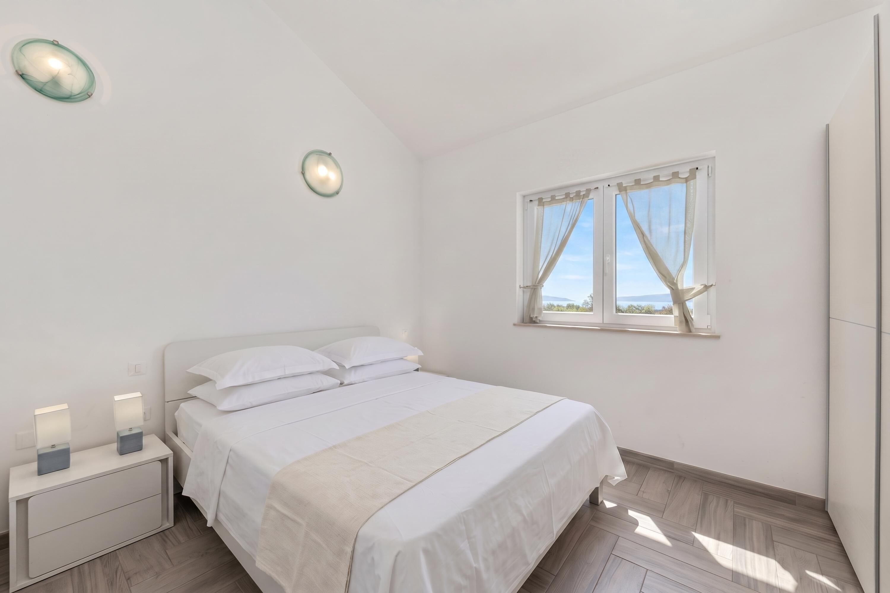 Un dormitorio con cama, mesitas de noche, una ventana con cortinas e iluminación en la pared.