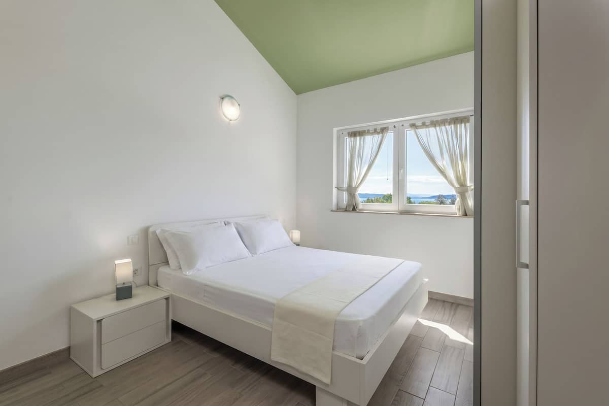 A bedroom with a bed, bedside tables, a window with curtains, and a view of the outdoors.