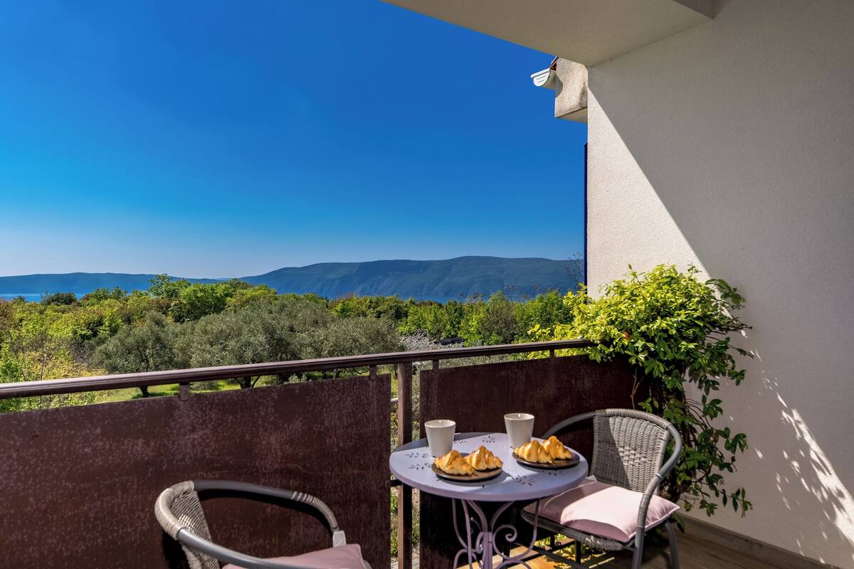A balcony with wicker chairs, a small round table, and a view of mountains and greenery.