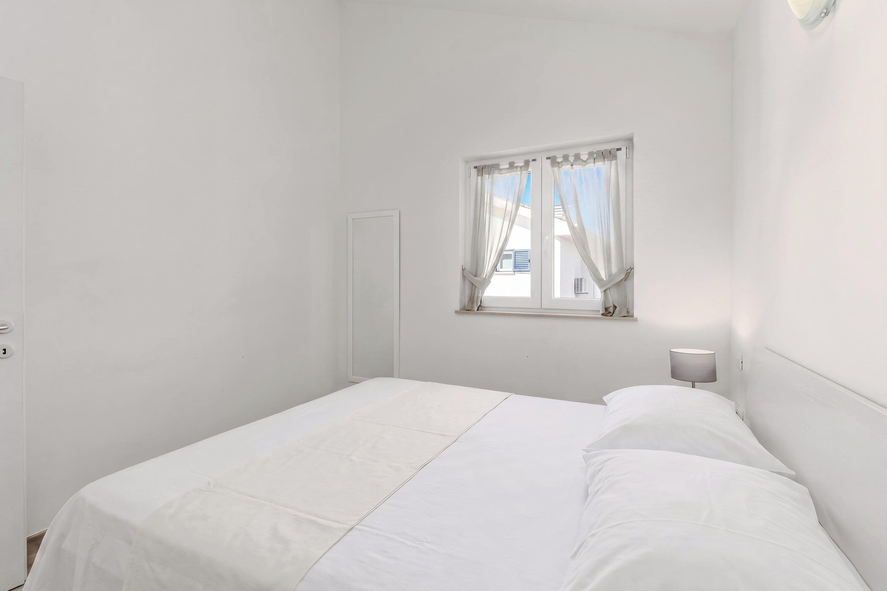 A simple, white room with a single bed, a window with curtains, and a wall-mounted light.