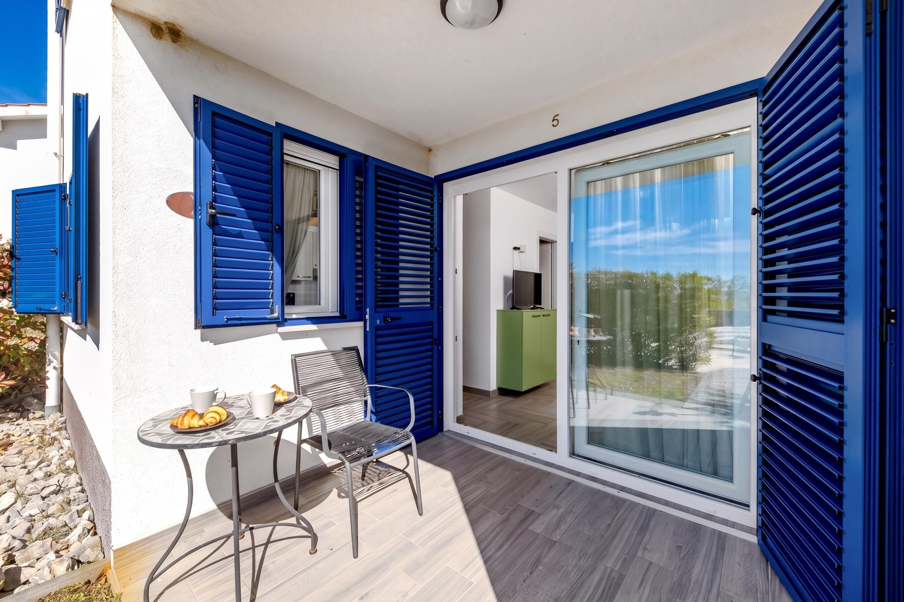 A balcony with a table, chairs, and a view of a green cabinet and a TV in the background.